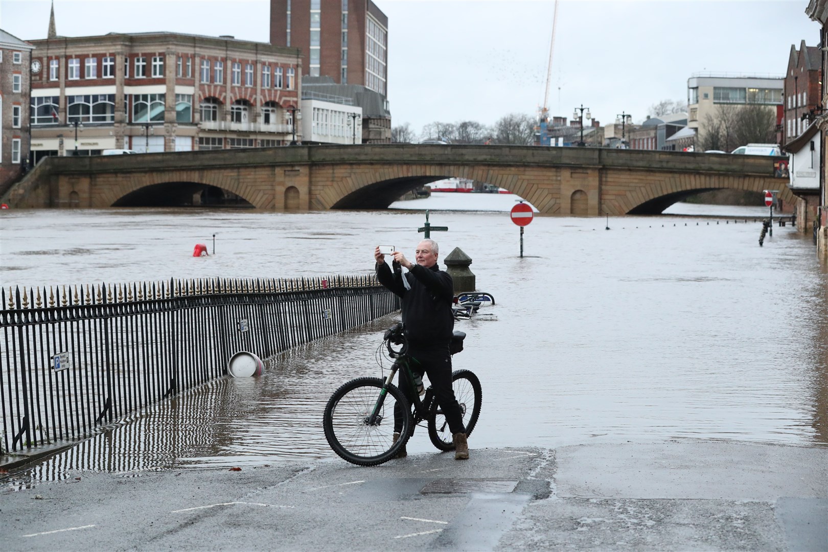 A man on a bicycle taking a photograph in front of floodwaters in York (Danny Lawson/PA)