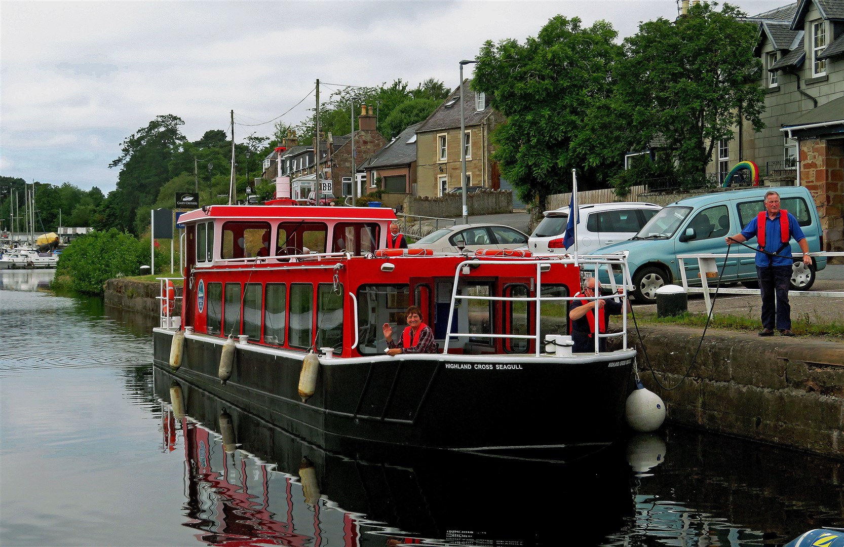 Highland Cross Seagull parked by the canalside, with volunteer crew members Brian Macleod on the towpath holding a mooring rope, and Kay Burton and Charlie Watt in the bow.